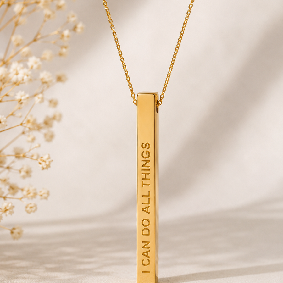 A gold bar necklace with the engraved text 'I CAN DO ALL THINGS' hanging from a delicate gold chain, styled with dried white flowers on a warm beige background.