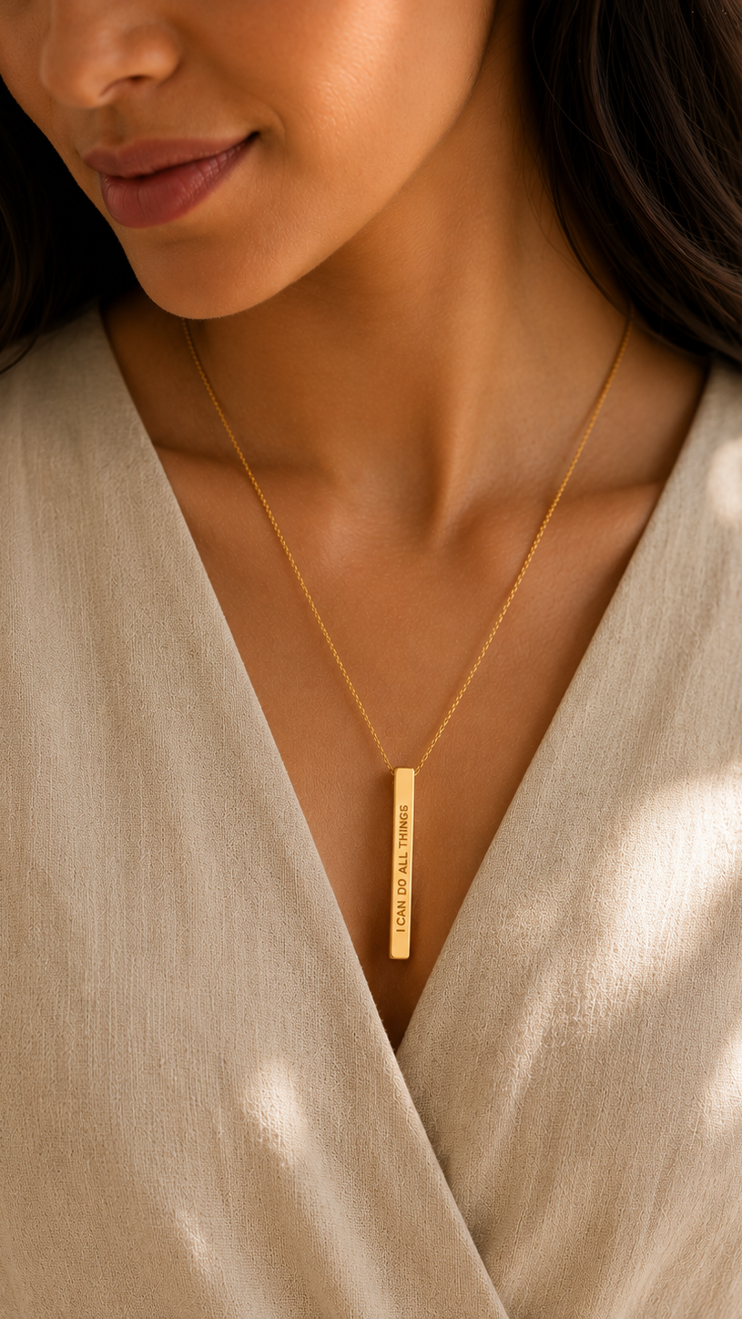 A close-up of a woman wearing a delicate gold chain necklace with a vertical bar pendant engraved with the words 'I CAN DO ALL THINGS', styled with a beige linen wrap top.