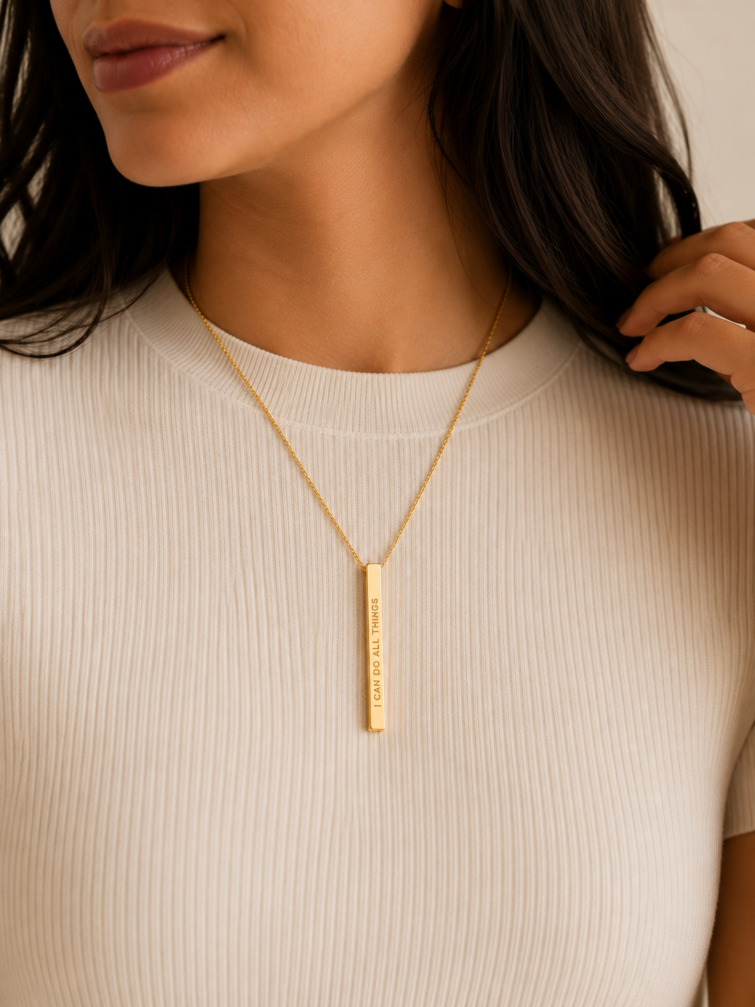 A close-up of a woman wearing a gold bar necklace with engraved text, paired with a cream ribbed top.