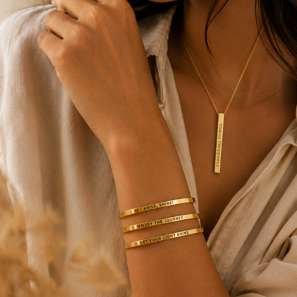A close-up lifestyle shot of a woman wearing three gold engraved bangles on her wrist and a matching gold bar pendant necklace. The bangles are engraved with 'BORN TO BE LOVED' and the necklace is engraved with 'BORN TO BE LOVED YOU'.