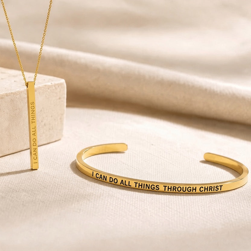 Gold jewelry set featuring a bar pendant necklace and a cuff bracelet with engraved text, displayed on a beige fabric background with a stone block prop.